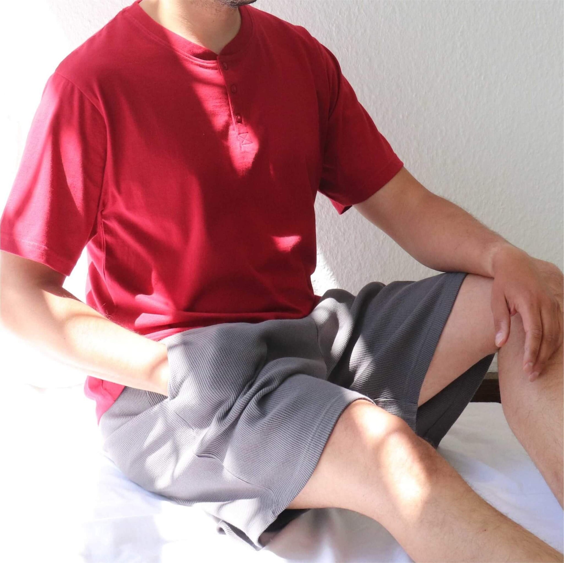 Man in rode shortama, bestaande uit T-shirt met knoopjes en grijze korte broek, zittend op witte ondergrond
